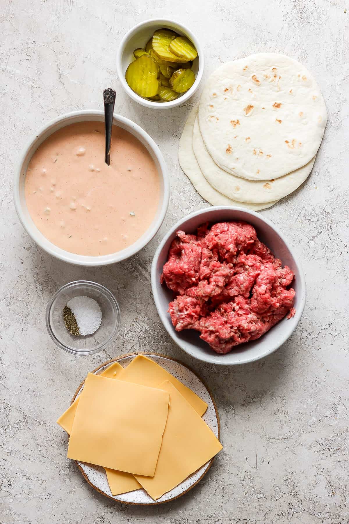 Individual bowls of burger sauce, ground beef, burger pickles, salt & pepper, and cheese.  Three flour corn tortillas are laying on the countertop next to the bowls.
