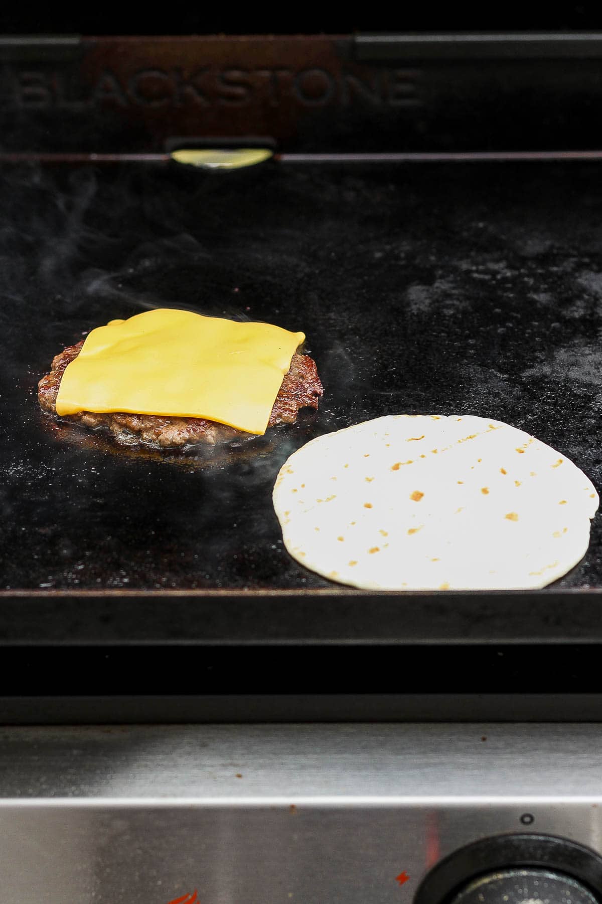 Burger patty topped with American cheese.  A tortilla is next to the burger patty on the blackstone.