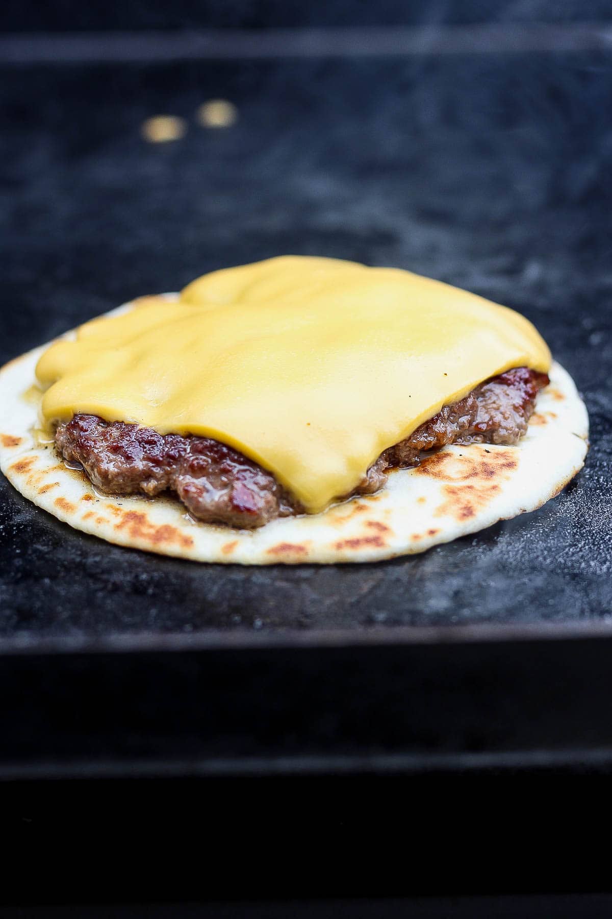The burger patty with cheese is on top of the tortilla on the blackstone.