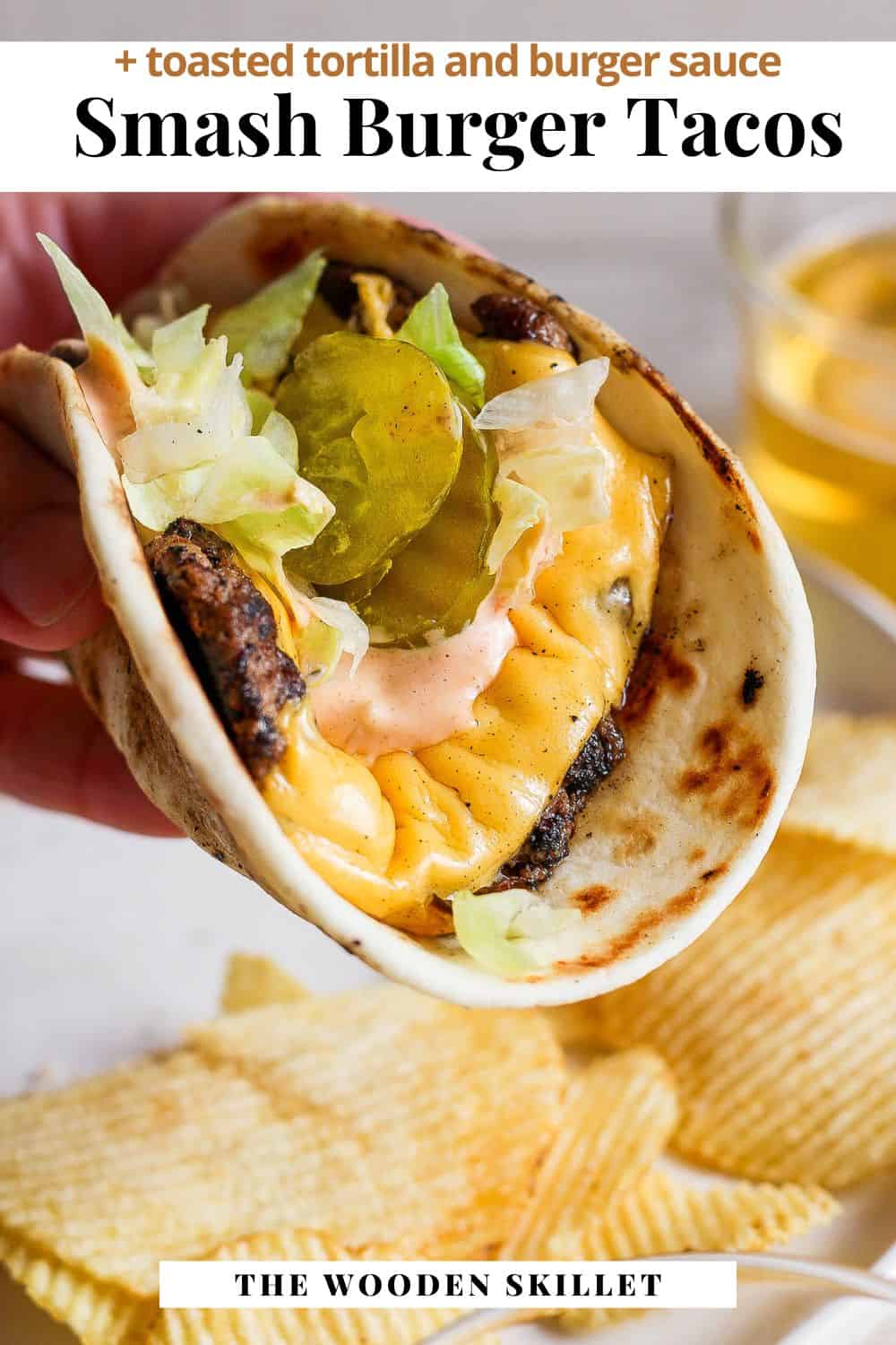 The smash burger taco being held like a taco above a plate of potato chips. Pinterest image title reads, "smash burger tacos + toasted tortilla and burger sauce"