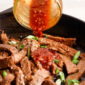 Someone pouring bulgogi sauce on top of some cooked bulgogi beef with green onions in a skillet.