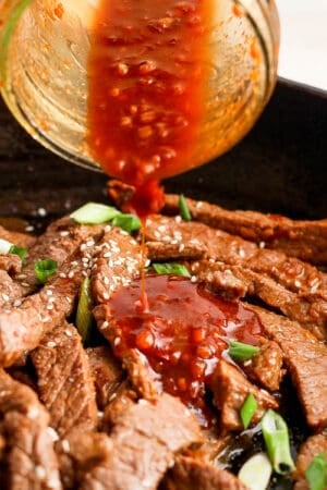 Someone pouring bulgogi sauce on top of some cooked bulgogi beef with green onions in a skillet.