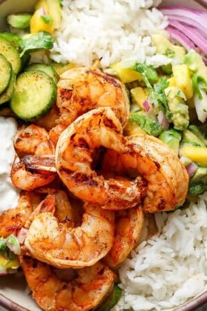 Top down shot of a bowl with rice, grilled chili lime shrimp, mango salsa, cucumbers and red onion.