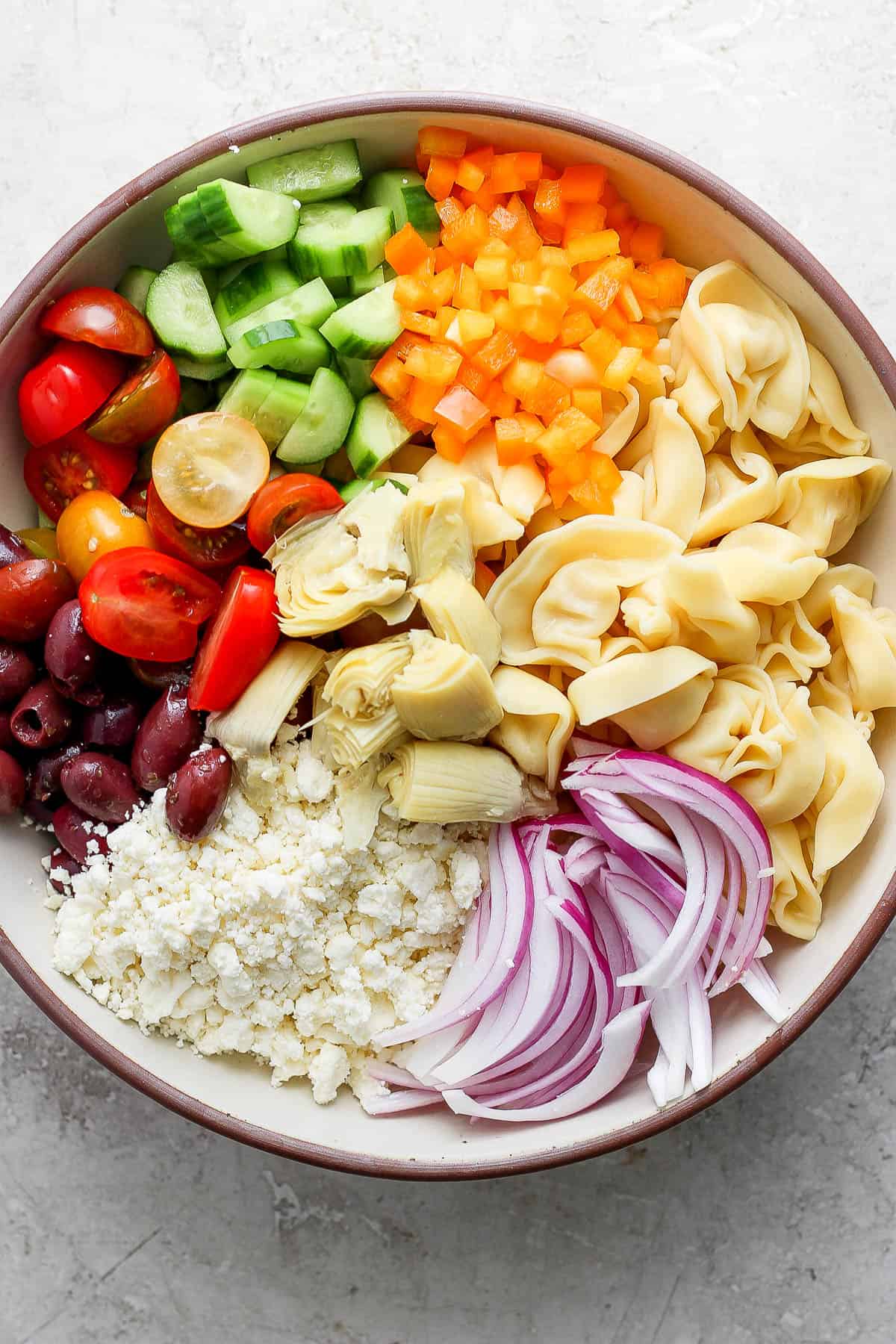 Feta cheese, sliced cherry tomatoes, sliced red onion, artichoke hearts, cheese tortellini, sliced cucumbers, olives and diced red bell pepper assembled in a bowl.