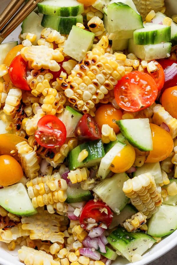 Top down shot of a bowl full of grilled corn salad with corn, tomato, cucumbers, and red onion with a spoon sticking out.