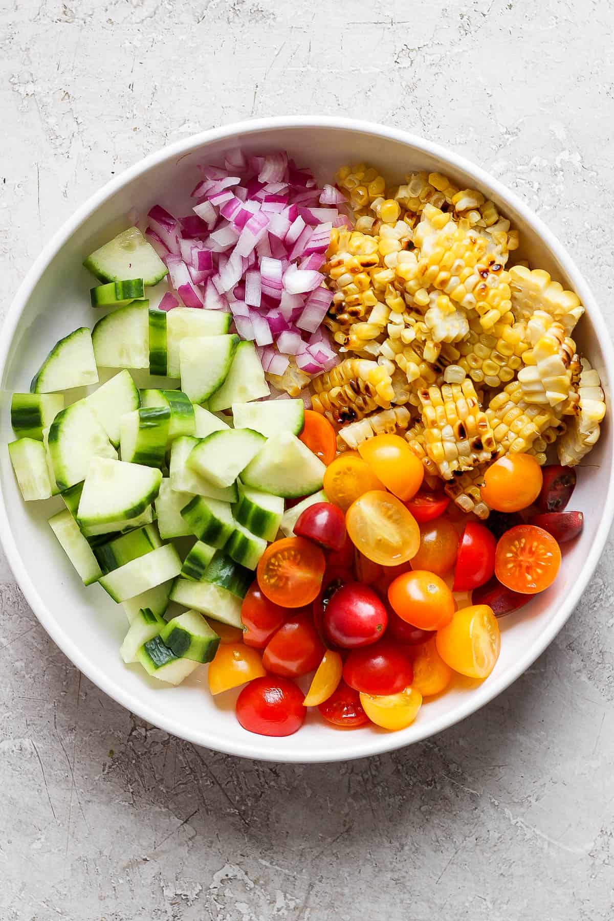 A large white bowl of cucumbers, red onion, tomatoes, and the corn cut off the cobs.