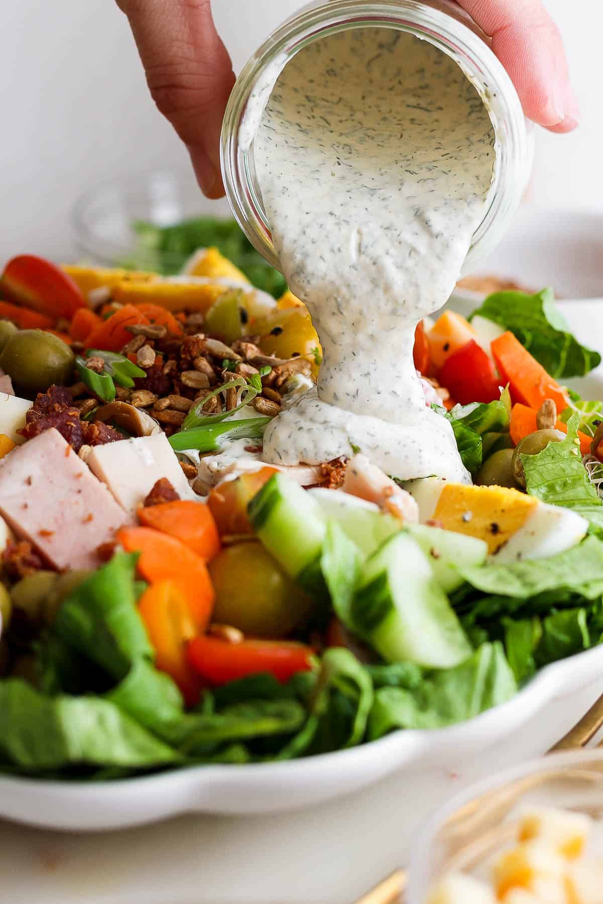 A hand pouring a jar of ranch dressing over a chef salad. 