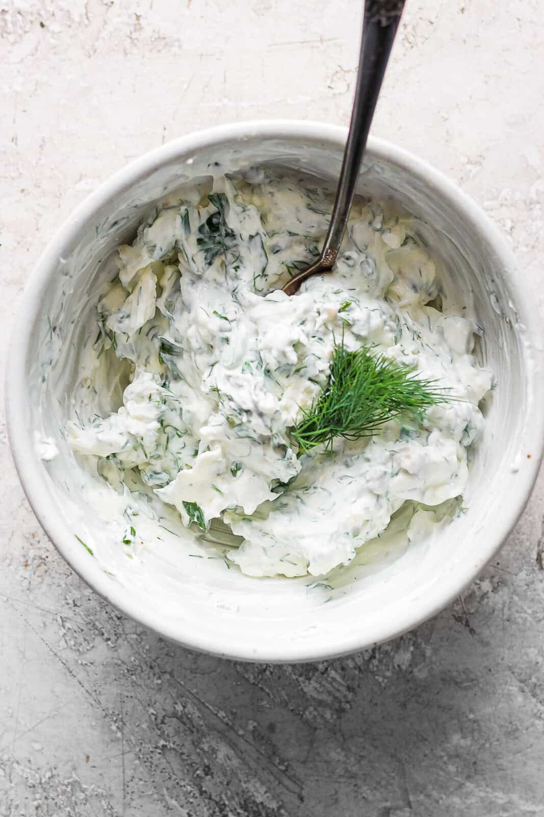 A bowl of chive and dill cream cheese sauce mixed in a bowl with fresh dill on top and a spoon sticking out. 