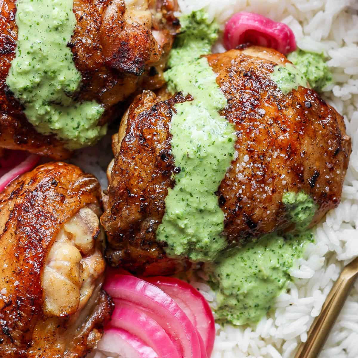 Top down shot of a grilled peruvian chicken thigh with peruvian green sauce on top on some white rice with pink quick pickled onions.