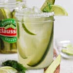 Straight on shot of a pickle margarita in a mason jar with a pickle inside and a jar of pickles behind it.