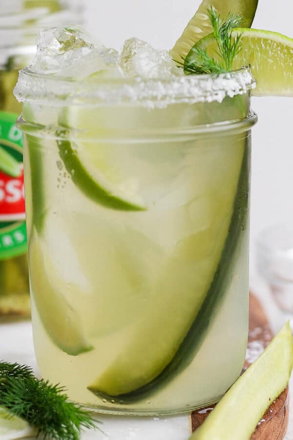 Straight on shot of a pickle margarita in a mason jar with a pickle inside and a jar of pickles behind it.
