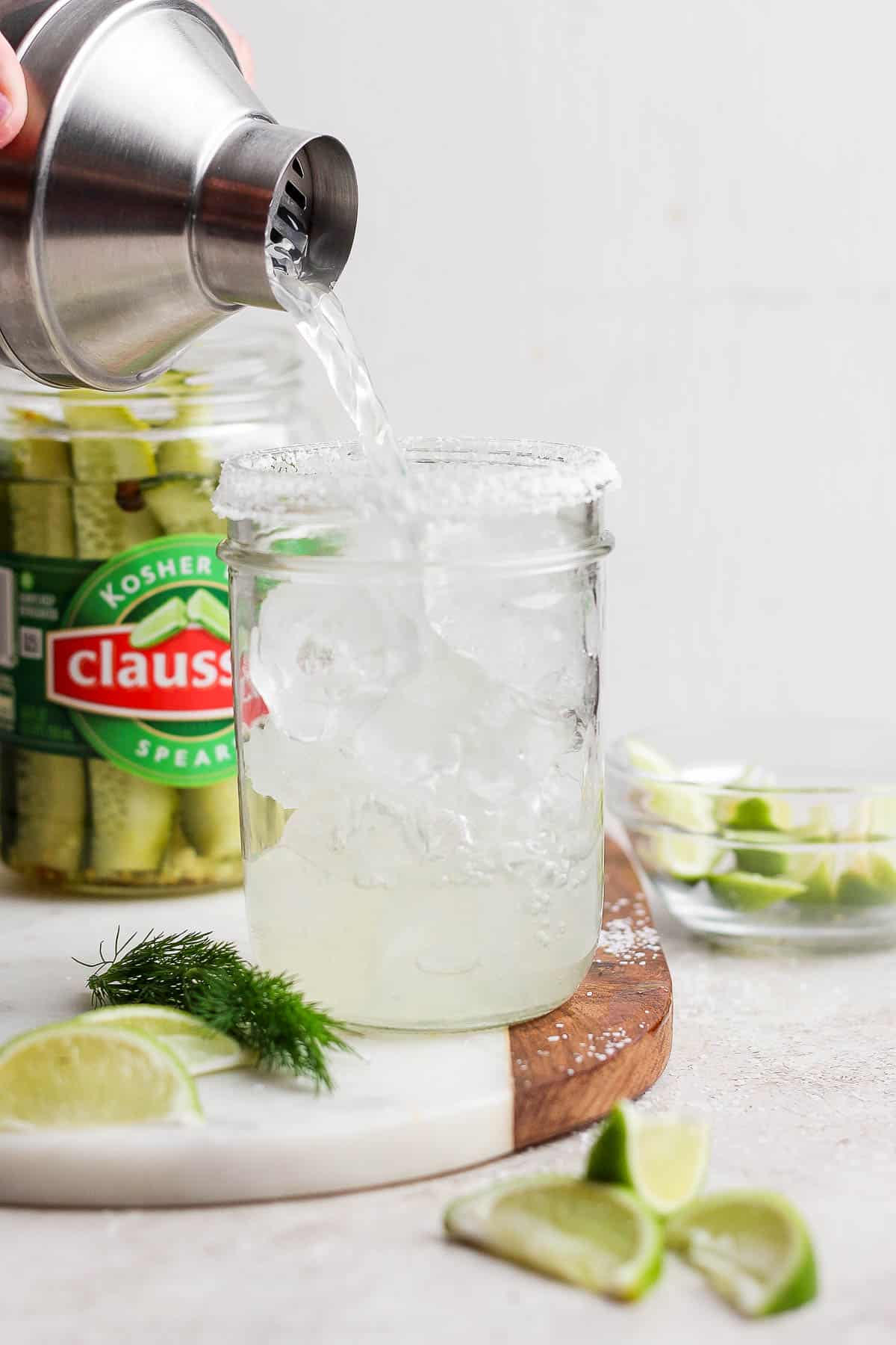 A dill pickle margarita being poured out of the shaker into a mason jar with ice.