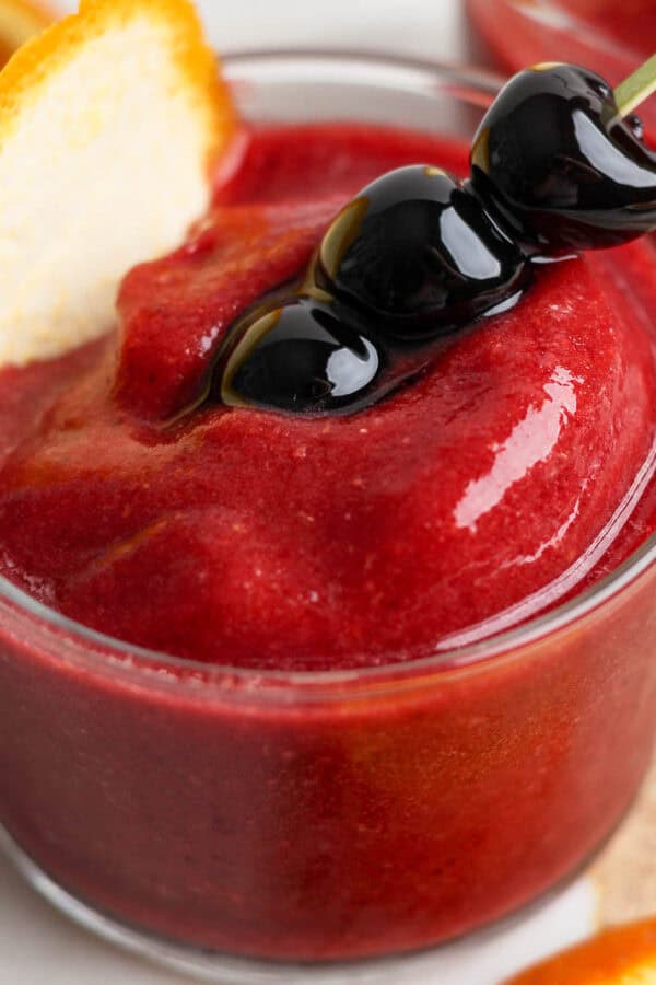 Side angle of a short glass filled with a frozen old fashioned and garnished with 3 cherries on a stick and orange peel.
