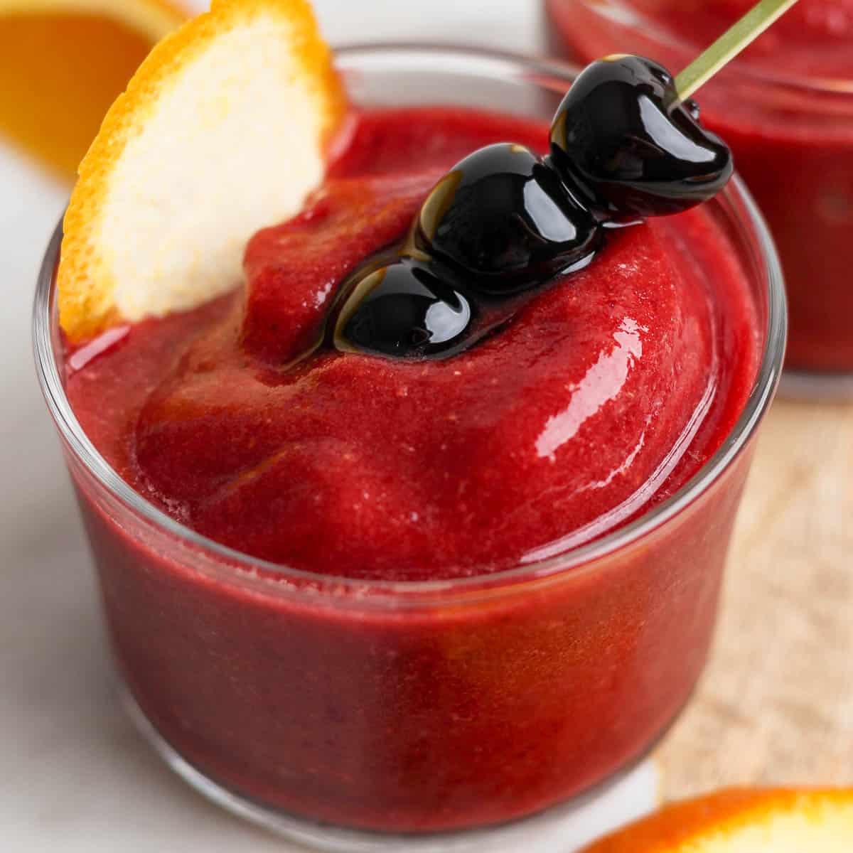 Side angle of a short glass filled with a frozen old fashioned and garnished with 3 cherries on a stick and orange peel.