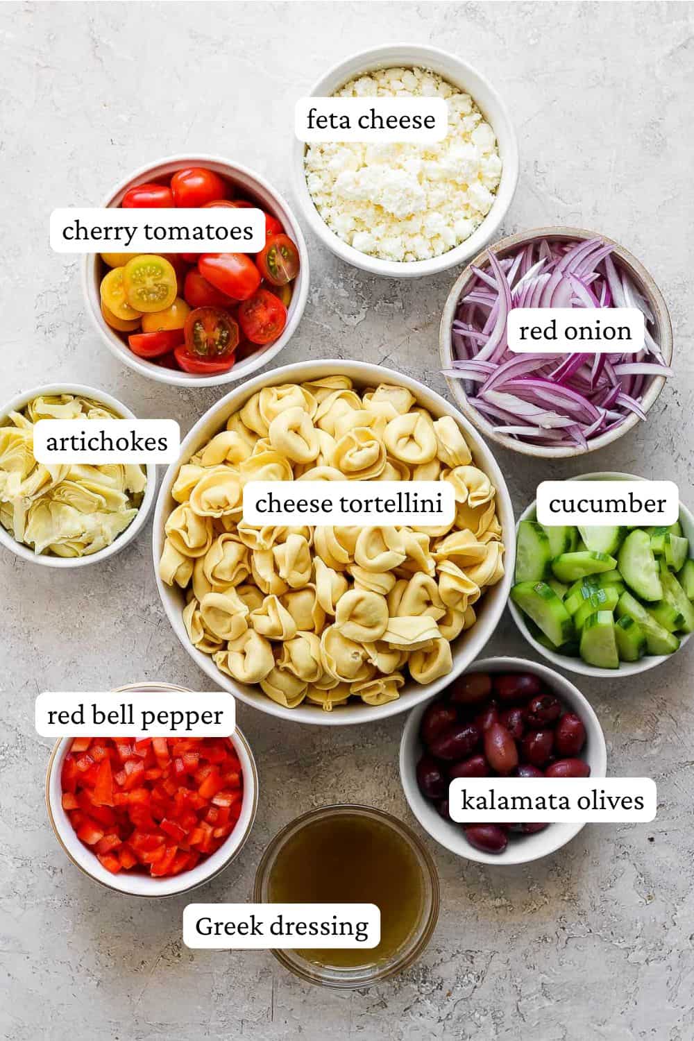 Greek tortellini salad ingredients in individual bowls including feta cheese, cherry tomatoes, red onion, artichokes, cheese tortellini, cucumber, red bell pepper, Kalamata olives, and Greek dressing.