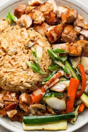 A plate of hibachi chicken with some vegetables and fried rice, green onion and yum yum sauce drizzled on top.