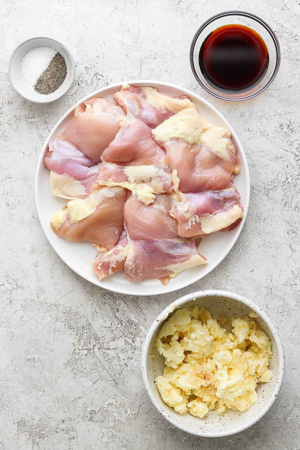 A plate of chicken thighs next to bowls of salt, pepper, soy sauce, and garlic butter.