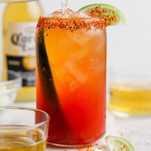 Straight on shot of a Michelada in a beer glass with Tajin rim and lime wedge with a Corona in the background.