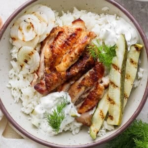 Top down shot of a bowl of rice with grilled pickle brined chicken thigh on top, sliced, with sauce, grilled pickles and grilled onion.