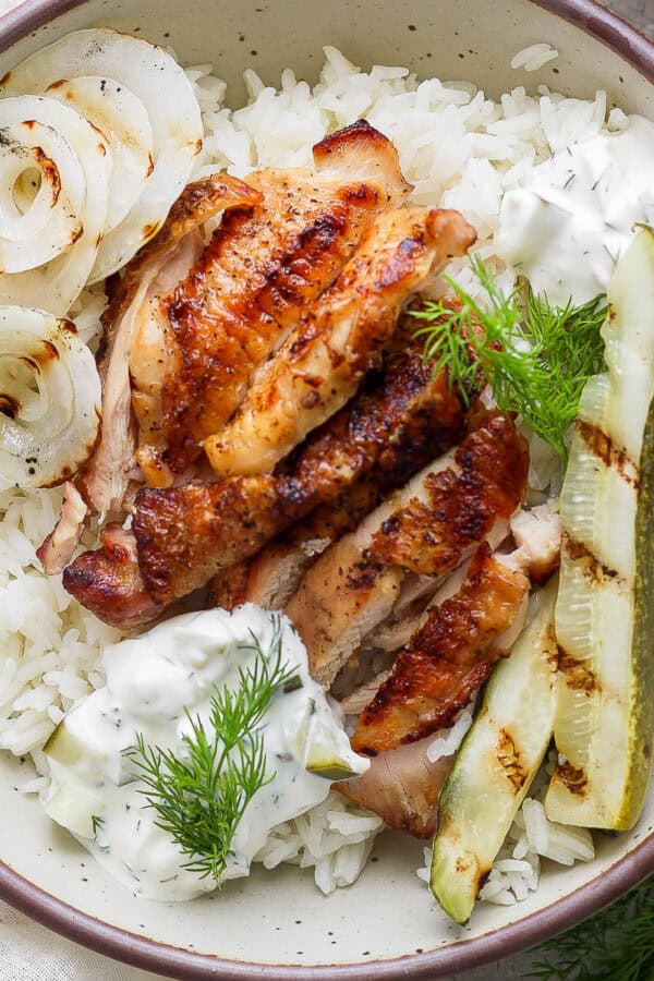 Top down shot of a bowl of rice with grilled pickle brined chicken thigh on top, sliced, with sauce, grilled pickles and grilled onion.