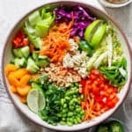 Top down shot of a bowl of ramen noodle salad with carrots, cabbage, cucumber, oranges, edamame and bell pepper.