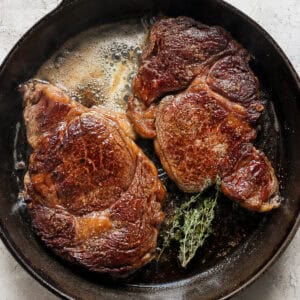 Top down shot of two ribeye steaks cooking in a cast iron skillet with melted butter and fresh thyme.