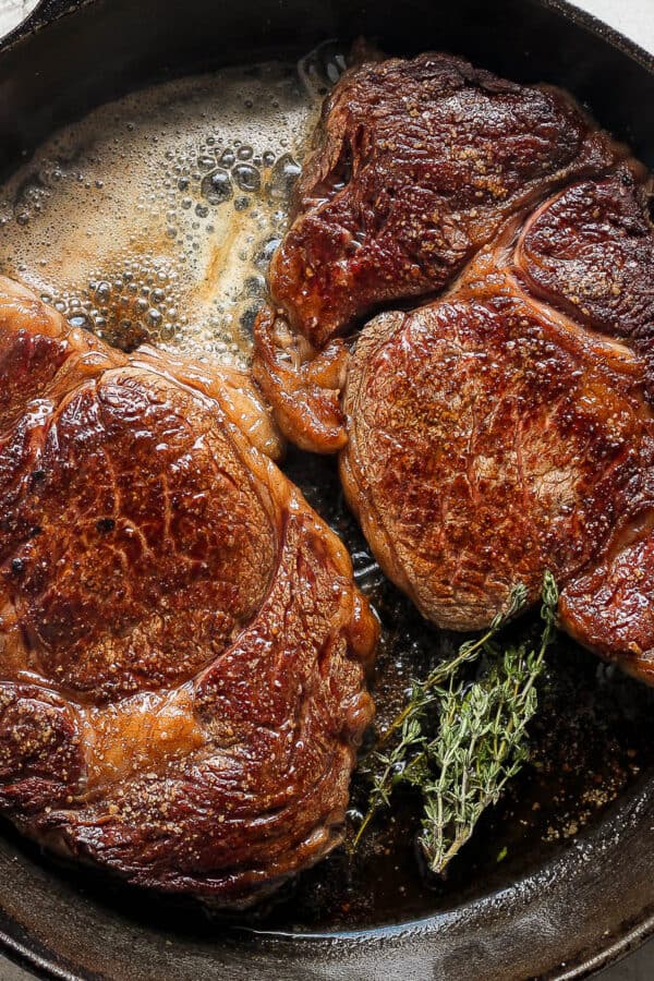 Top down shot of two ribeye steaks cooking in a cast iron skillet with melted butter and fresh thyme.
