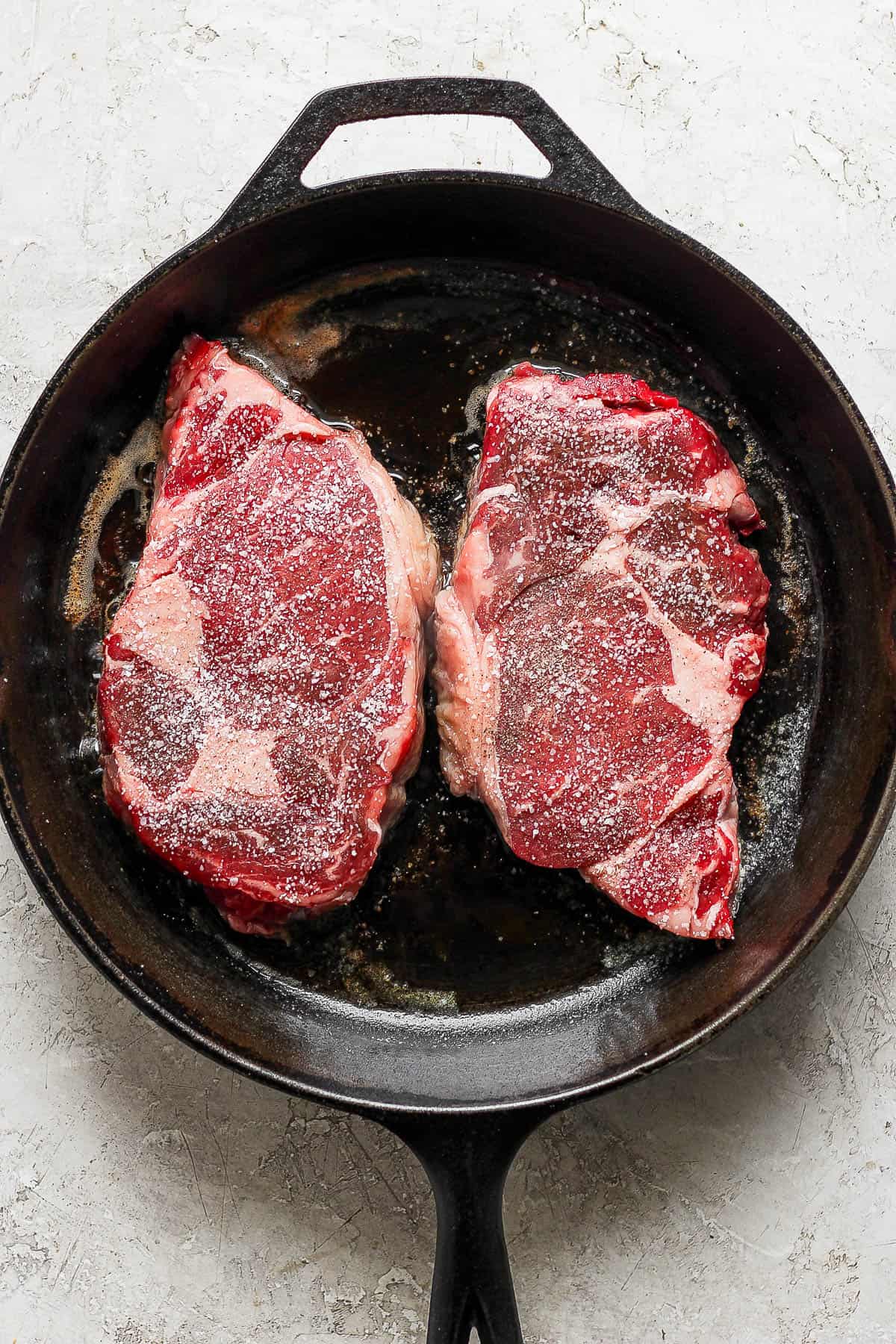Two ribeye steaks seasoned with salt and pepper placed in a hot cast iron skillet to sear.