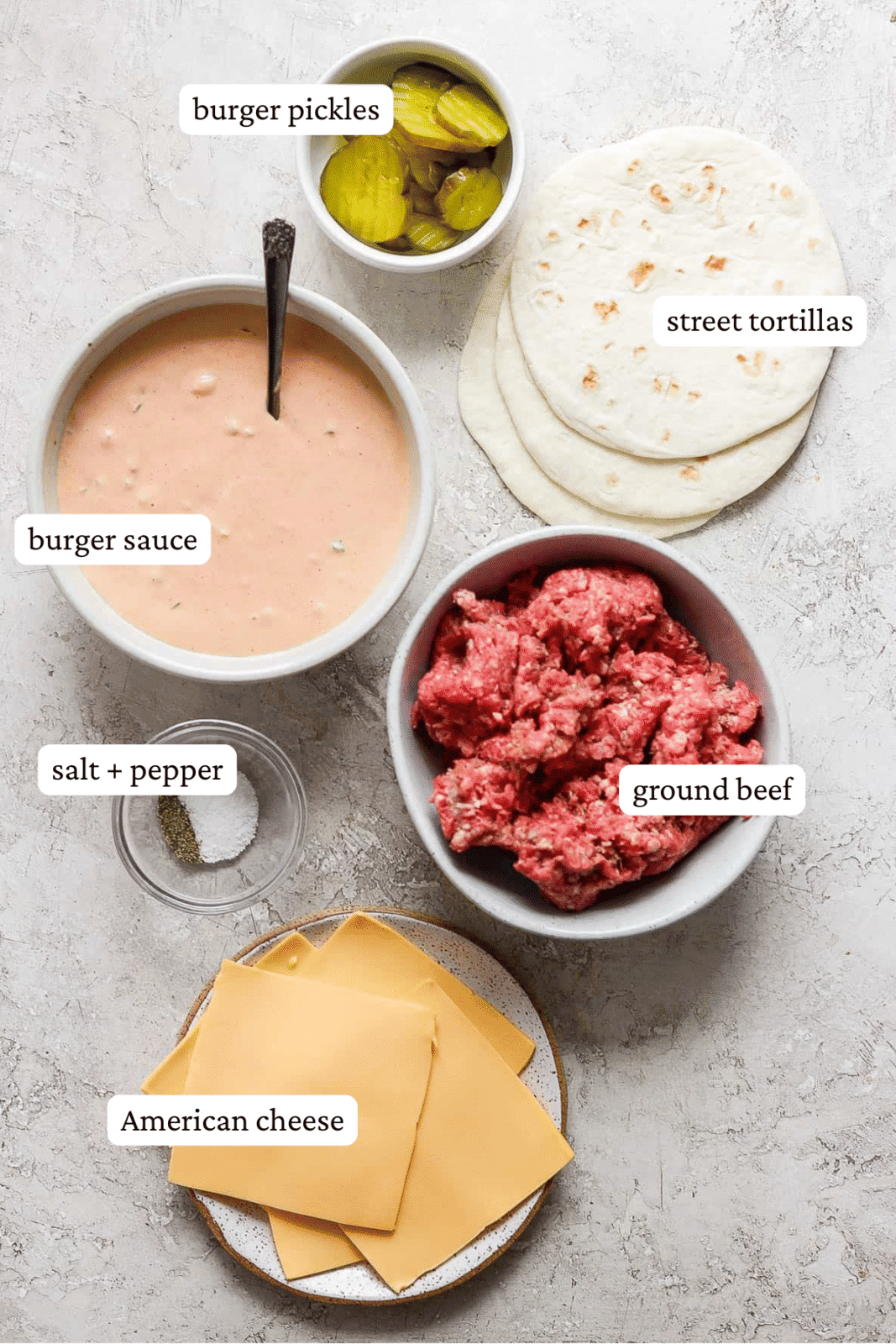Individual bowls of burger sauce, ground beef, burger pickles, salt & pepper, and cheese. Three flour corn tortillas are laying on the countertop next to the bowls.