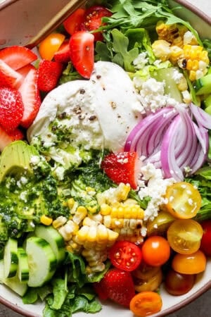 Top down shot of a big bowl of summer salad with grilled corn, burrata, basil vinaigrette, strawberries, avocado and tomatoes.