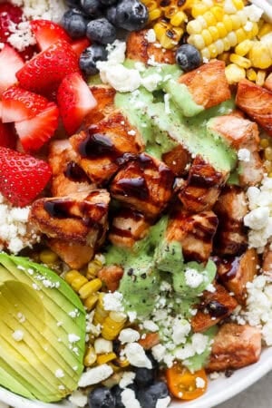 Bowl filled with salmon bites with balsamic glaze, avocado, feta cheese, strawberries, blueberries, grilled corn and a creamy basil sauce.