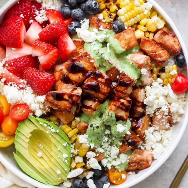 Bowl filled with salmon bites with balsamic glaze, avocado, feta cheese, strawberries, blueberries, grilled corn and a creamy basil sauce.