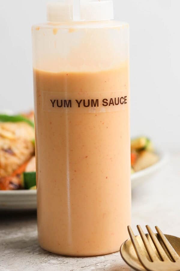 A tall plastic bottle of yum yum sauce with a label on it with a plate of hibachi chicken behind it and a fork and spoon laying in front of it.