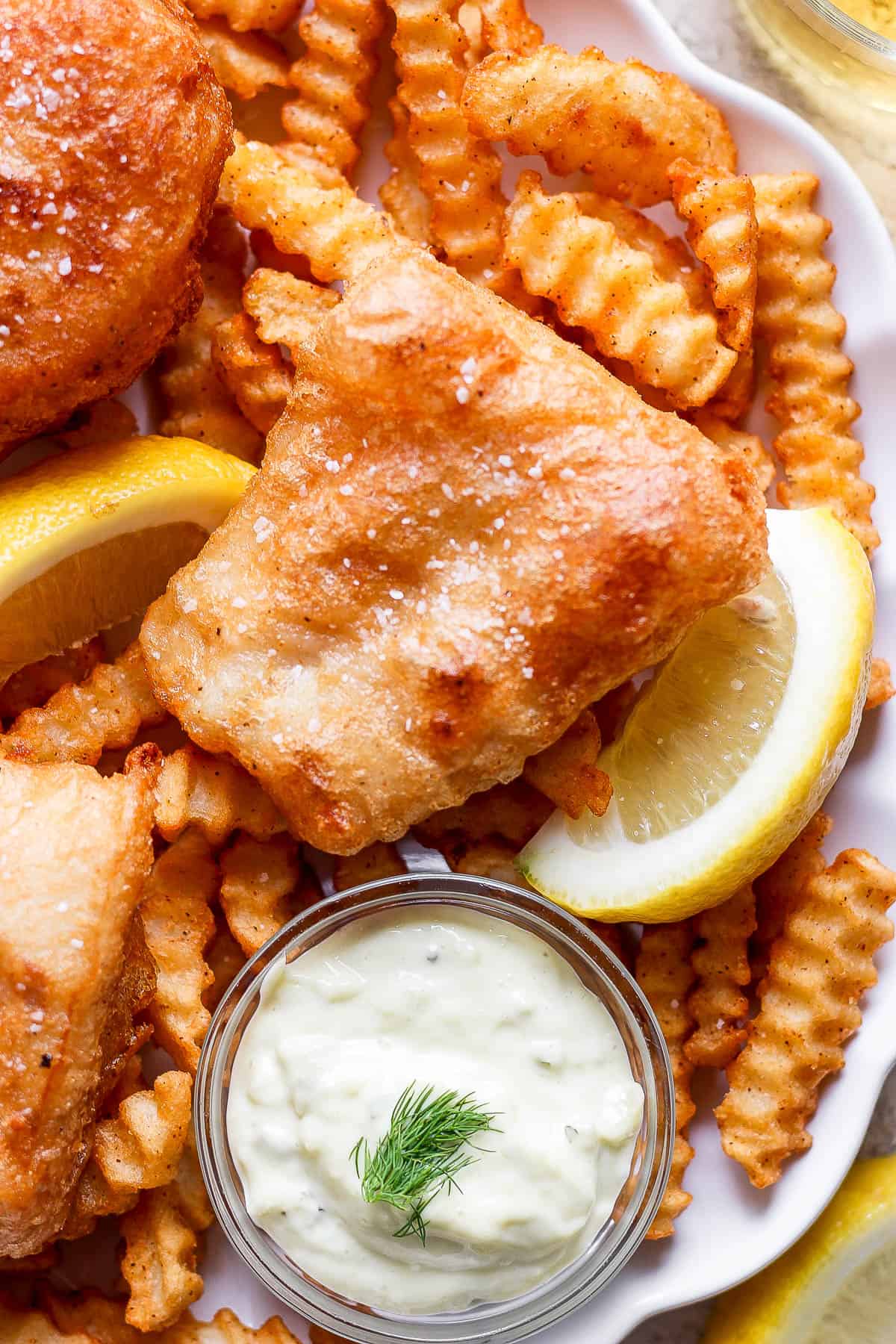 Beer battered fish on a plate with french fries, tartar sauce, and lemon wedges.