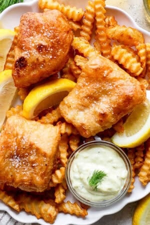 Top down shot of a plate of french fries with beer battered fish on top with fresh lemon wedges and a little bowl of homemade tartar sauce.