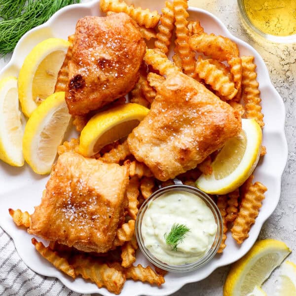 Top down shot of a plate of french fries with beer battered fish on top with fresh lemon wedges and a little bowl of homemade tartar sauce.