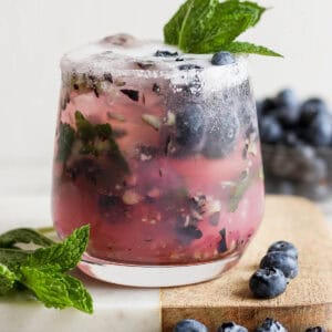 Straight on shot of a glass filled with a blueberry mojito on a marble board with fresh blueberries and fresh mint garnish.