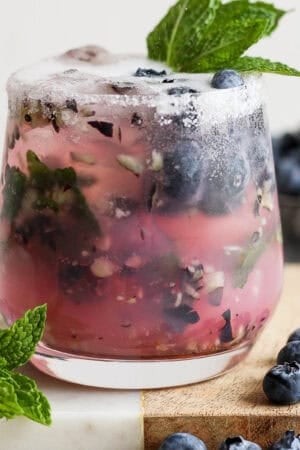 Straight on shot of a glass filled with a blueberry mojito on a marble board with fresh blueberries and fresh mint garnish.
