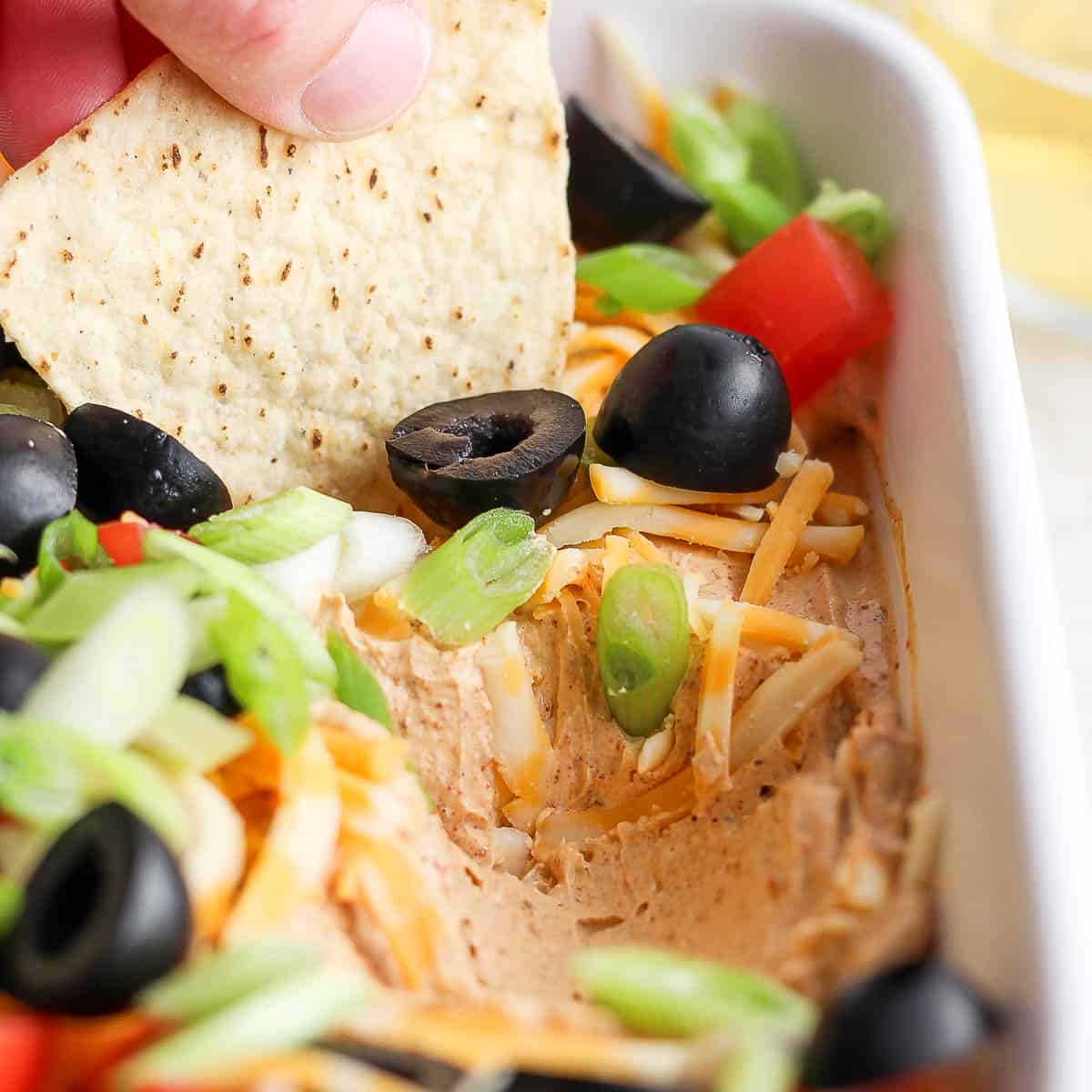 Side angle shot of someone using a tortilla chip to scoop out some taco dip from a casserole dish.