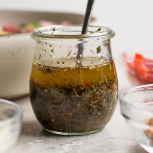 Straight on shot of a small jar of homemade italian dressing with a spoon sticking out and a bowl of salad behind it.