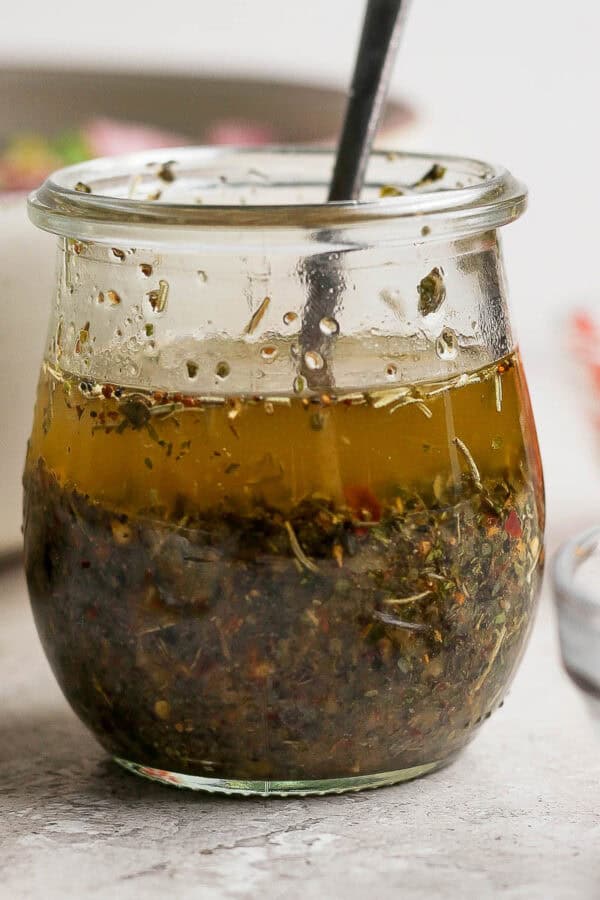 Straight on shot of a small jar of homemade italian dressing with a spoon sticking out and a bowl of salad behind it.