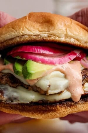 Someone holding a double-stacked turkey smash burger on a bun with burger sauce, avocado and quick-pickled onions.