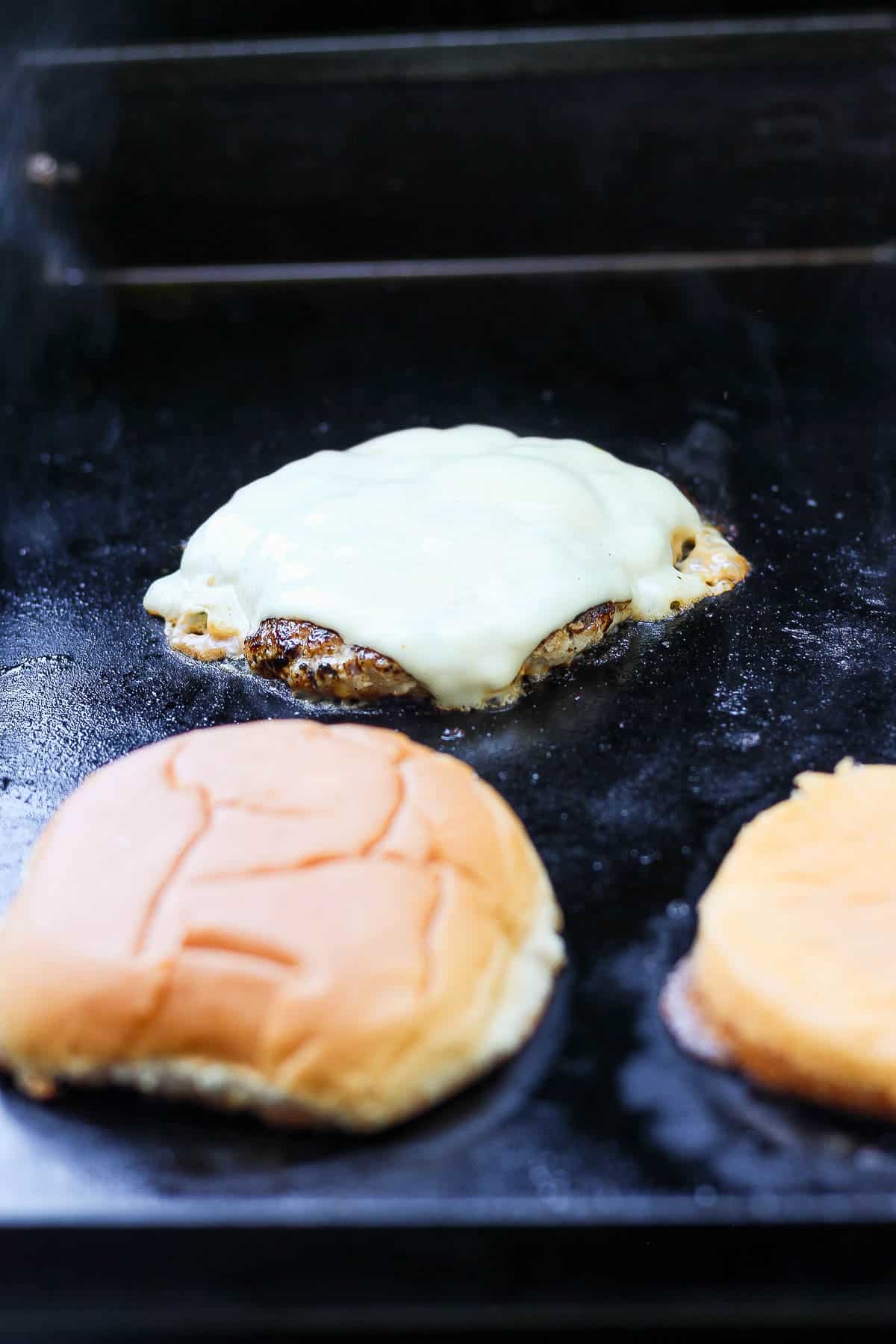 Burger patty with a slice of cheese on top next to a burger bun on the griddle.