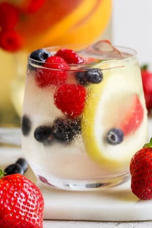 Straight on shot of a glass of white sangria with lemon, raspberries and blueberries.