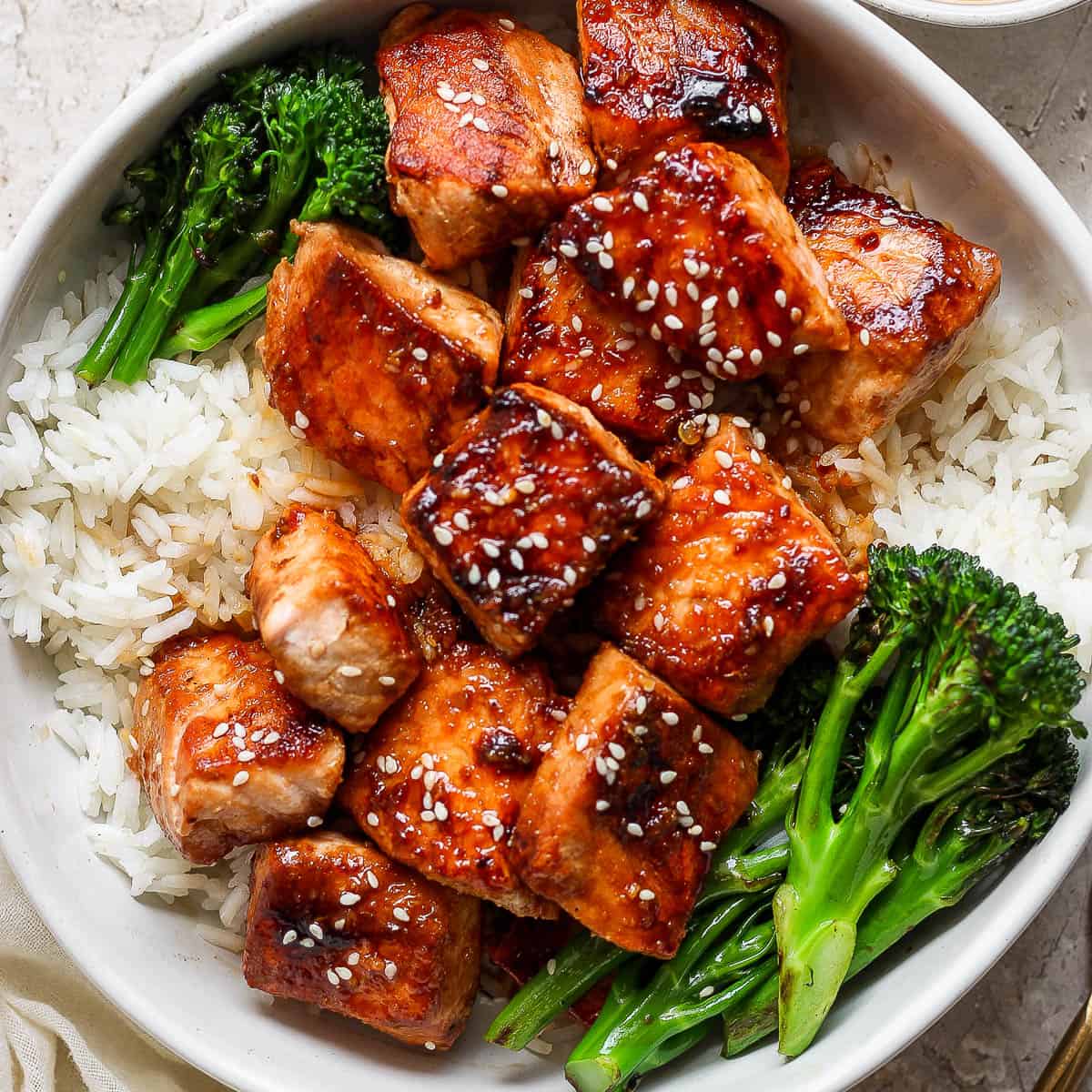 Top down shot of a bowl of rice with broccolini on the sides with cooked salmon bites in the middle with sesame seeds on top.