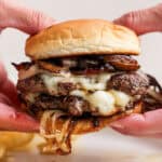 Someone holding a double-stacked mushroom Swiss burger recipe with melted Swiss cheese and onions falling out.