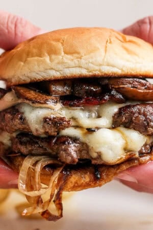 Someone holding a double-stacked mushroom Swiss burger recipe with melted Swiss cheese and onions falling out.