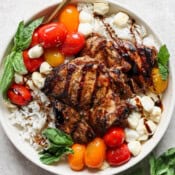 Grilled balsamic chicken thighs, cherry tomatoes, mozzarella balls, fresh basil, and balsamic glaze on rice in a white bowl.