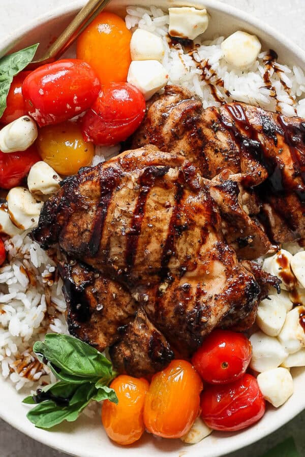 Grilled balsamic chicken thighs, cherry tomatoes, mozzarella balls, fresh basil, and balsamic glaze on rice in a white bowl.