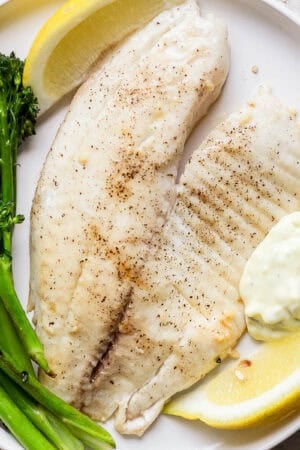 A plate with a piece of grilled tilapia, broccolini, lemon wedges and tartar sauce.